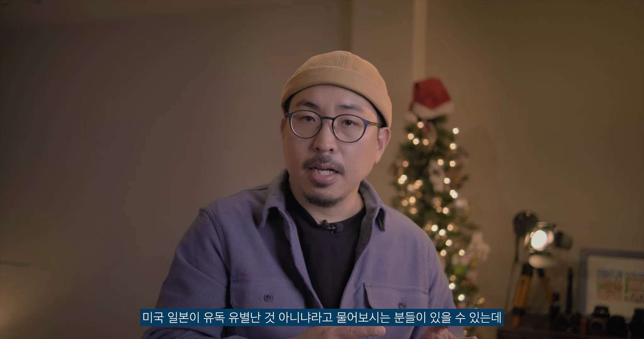 노란색 비니와 안경을 쓴 남성이 실내에서 카메라를 응시하며 이야기하고 있으며, 뒤편에는 크리스마스 트리가 장식되어 있는 영상 화면