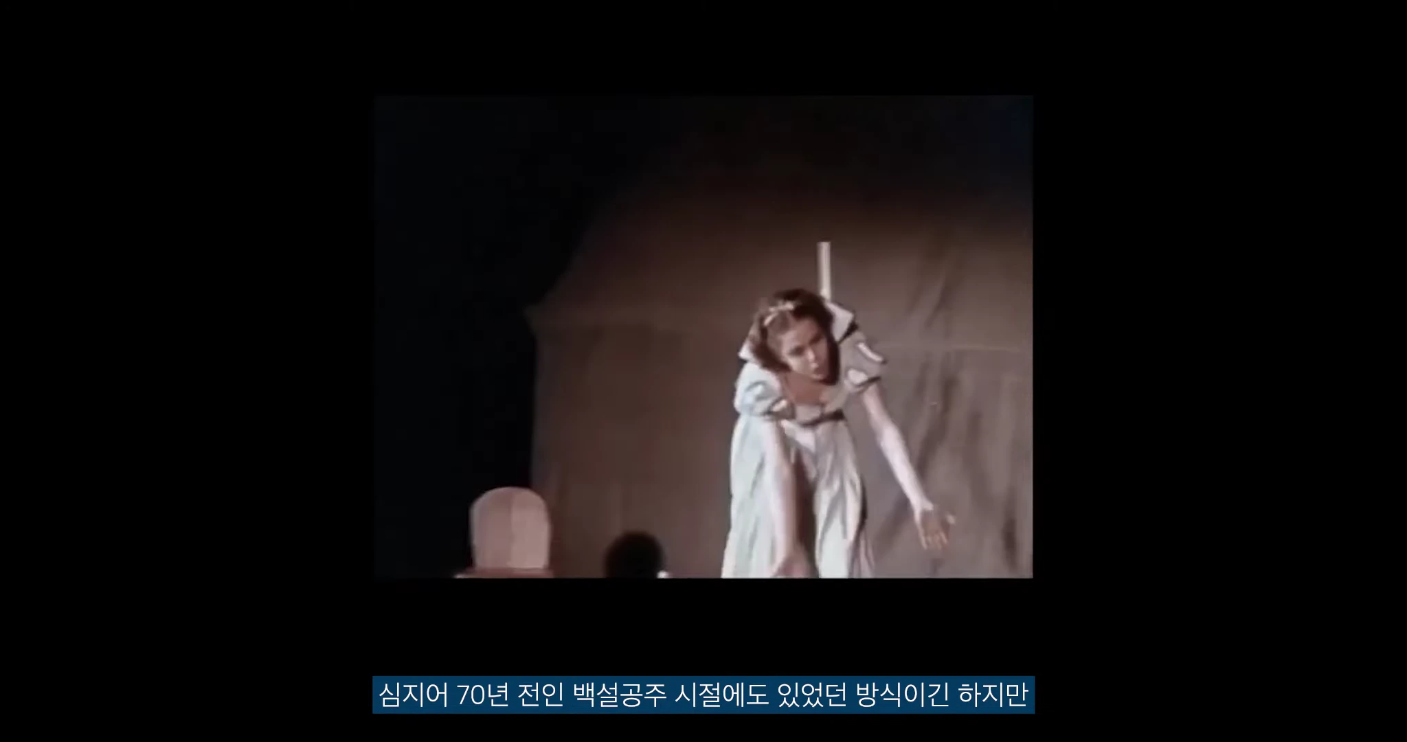 옛날 애니메이션 영화 속 한 장면으로, 드레스를 입은 여성이 조명 아래에서 손을 뻗으며 연기하는 모습이 흑백 화면에 담겨 있다.
