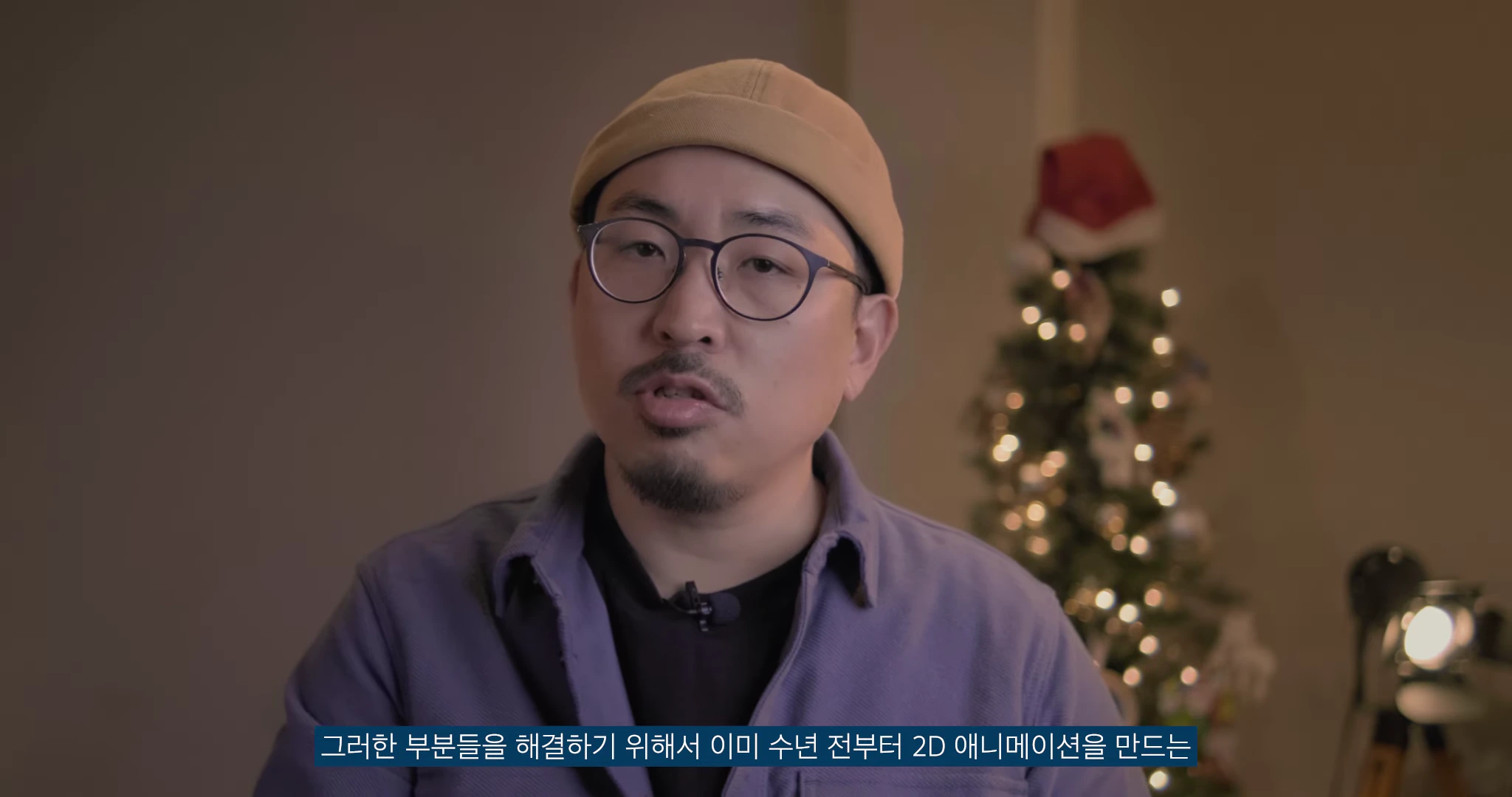 비니를 쓰고 안경을 쓴 남성이 정면을 바라보며 이야기하고 있으며, 배경에는 조명이 켜진 크리스마스 트리가 희미하게 보인다.