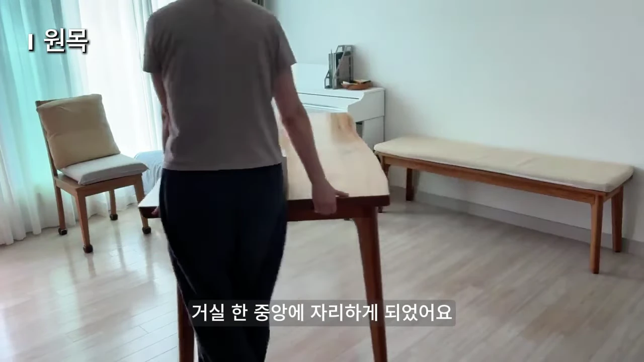 밝은 톤의 거실 한가운데에 놓인 커다란 원목 식탁을 한 사람이 옆에서 잡고 있는 모습입니다. 화면 상단에는 '1 원목'이라는 자막이, 하단에는 '거실 한 중앙에 자리하게 되었어요'라는 자막이 보입니다.