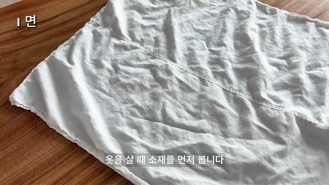 나무 테이블 위에 펼쳐진 구김이 있는 흰색 면 소재의 천 조각과 '면'이라는 글자가 적힌 영상 화면