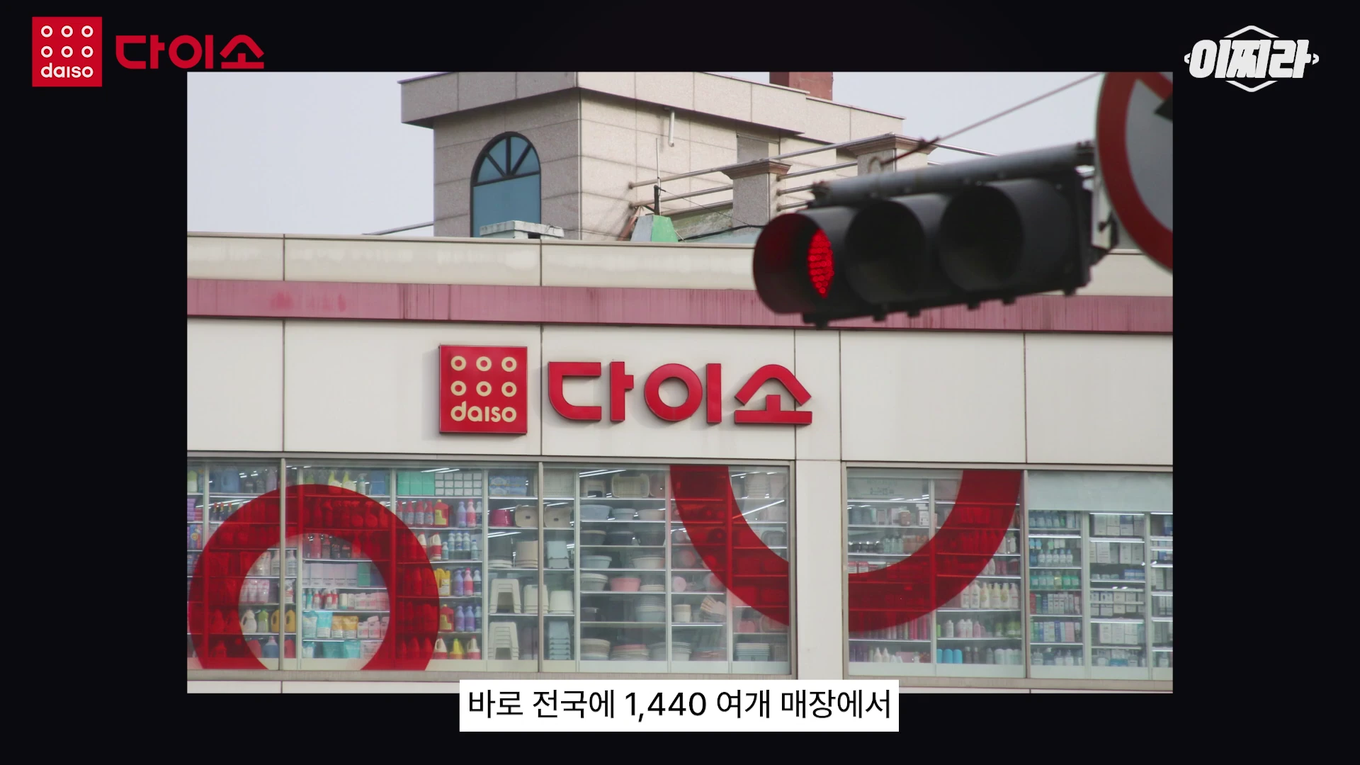 붉은색 다이소 로고가 부착된 매장 외관의 모습이며, 통유리창 너머로 진열된 상품들이 보이고 우측 상단에는 신호등이 위치해 있습니다.