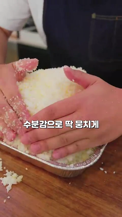 요리사가 앞치마를 입고 양손을 사용해 금속 용기 안에 든 닭 위로 소금을 두껍게 덮어 다지고 있는 모습