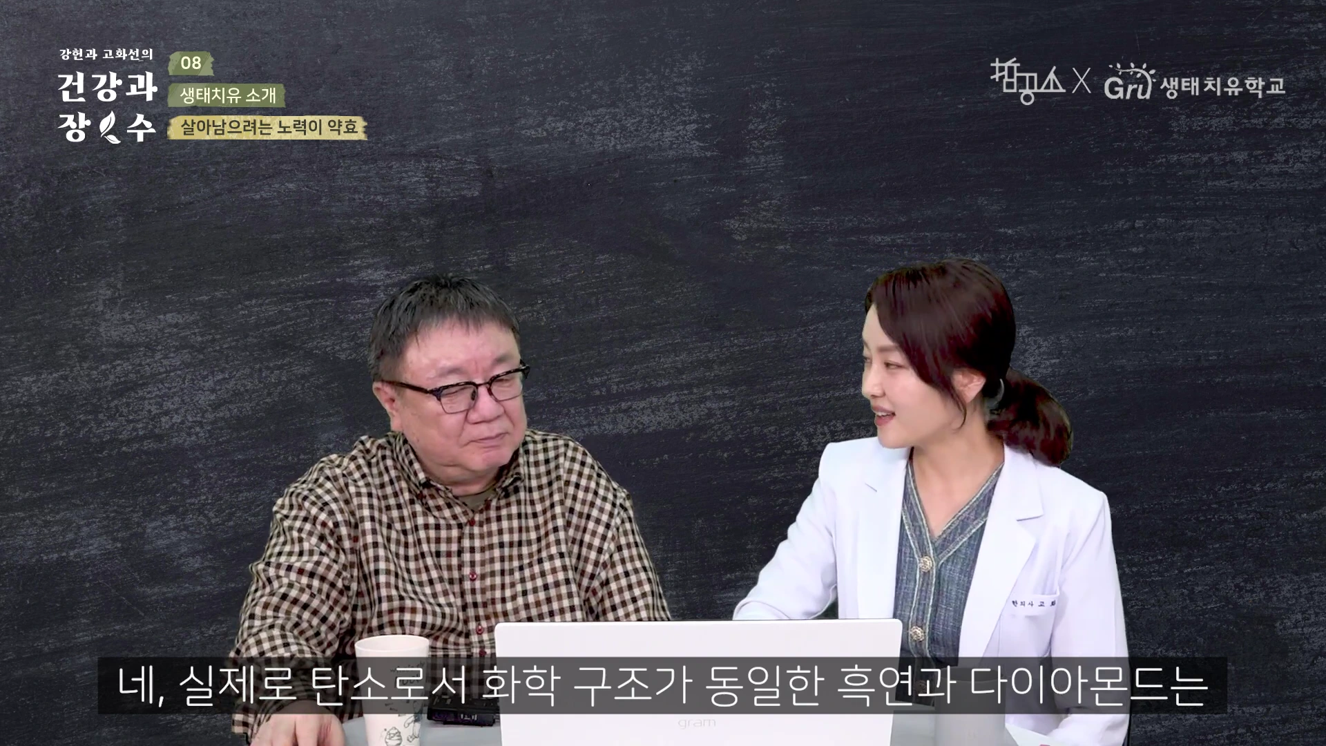 체크무늬 셔츠를 입은 남성과 흰색 의사 가운을 입은 여성이 책상을 사이에 두고 대화를 나누고 있는 스튜디오 인터뷰 장면이며 화면 하단에는 자막이 떠 있습니다.