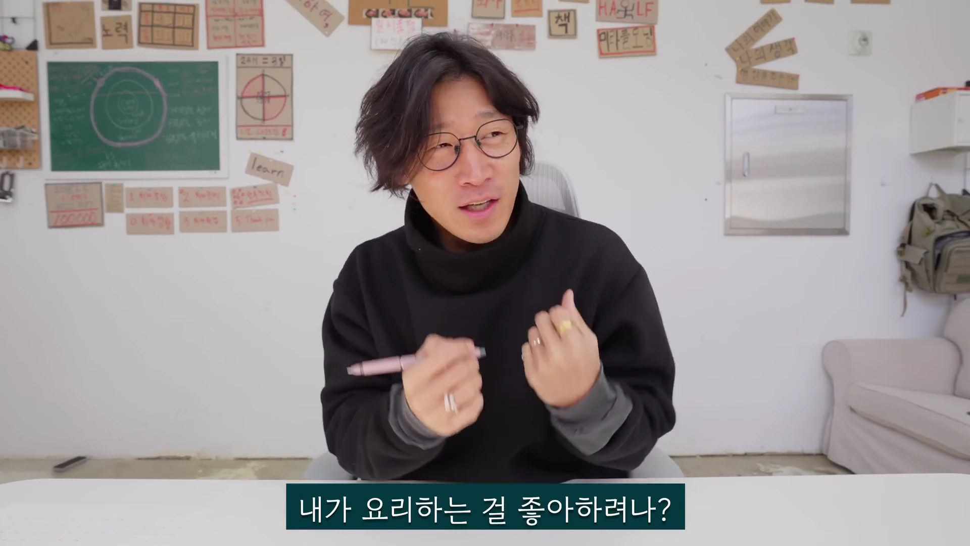 검은색 터틀넥을 입고 안경을 쓴 남성이 사무실 책상에 앉아 손동작을 하며 이야기하고 있다. 배경에는 메모지들이 붙은 흰 벽과 칠판이 보인다.