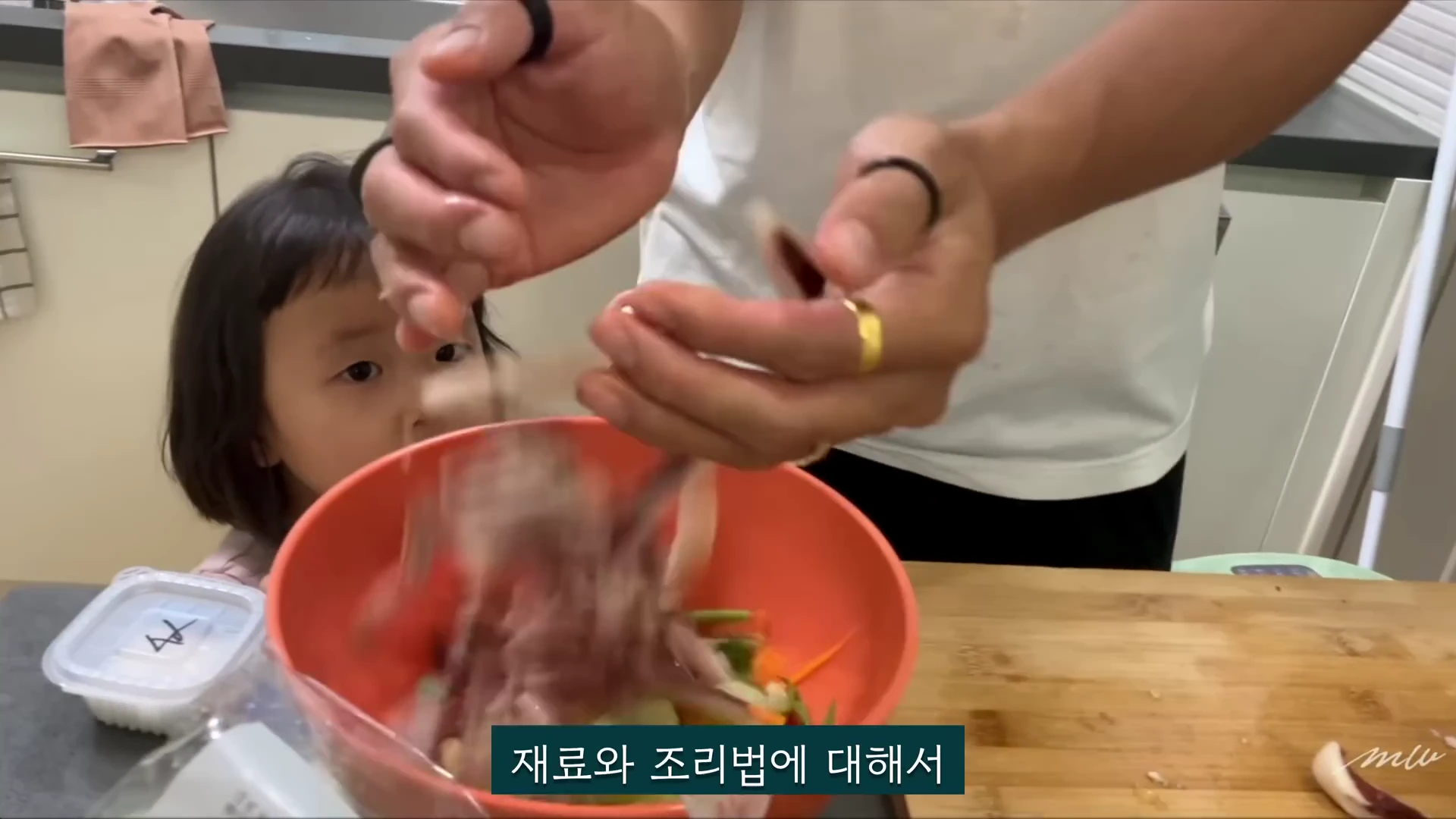 부엌에서 요리하는 남성의 손과 그 모습을 지켜보는 어린아이가 찍힌 영상 화면.