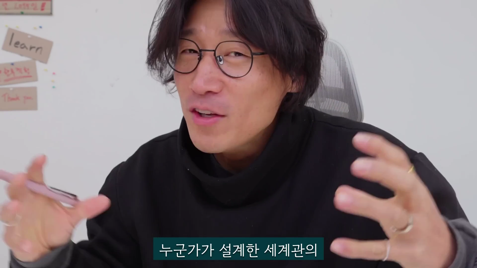 안경을 쓴 남성이 검은색 터틀넥을 입고 사무실 환경에서 손동작을 하며 이야기를 나누고 있으며, 화면 하단에는 자막이 떠 있고 배경 벽에는 메모들이 붙어 있다.