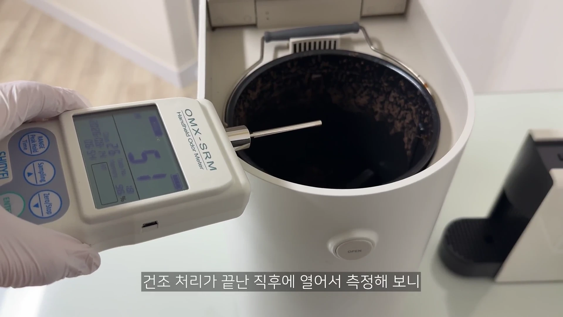 흰색 장갑을 낀 손이 디지털 냄새 측정기를 들고 음식물 처리기 내부의 냄새 수치를 확인하는 근접 촬영 사진