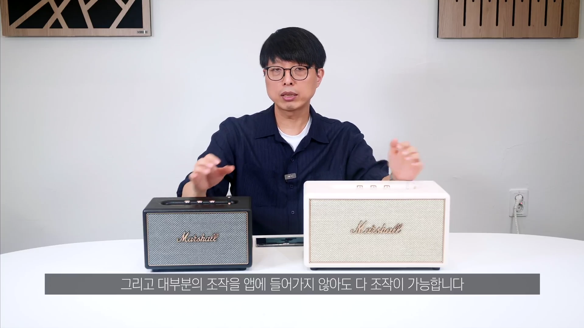 안경을 쓴 남성이 테이블 위에 놓인 검은색과 크림색 마샬 스피커 두 대를 앞에 두고 설명하고 있다.
