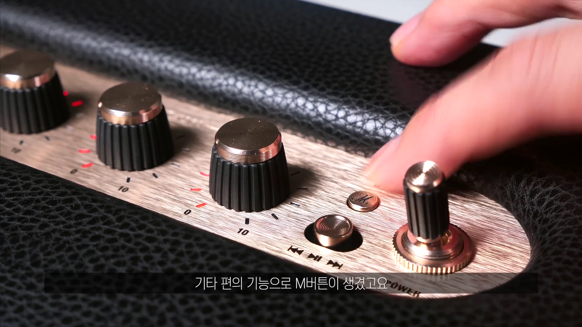 마샬 스피커 상단 패널의 금속 조절 다이얼과 버튼들을 손가락으로 조작하는 근접 촬영 장면입니다.