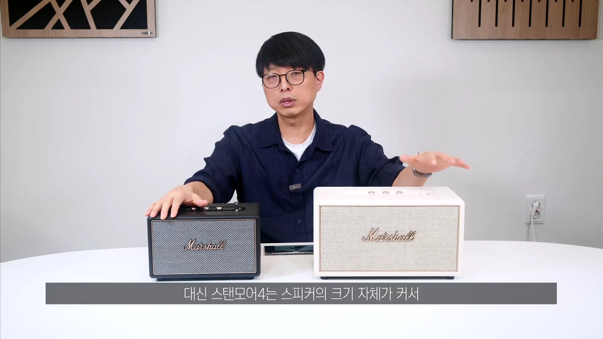 안경을 쓴 남성이 테이블 위에 놓인 검은색과 흰색의 마샬 스피커 두 대를 양손으로 가리키며 설명하는 영상 장면.