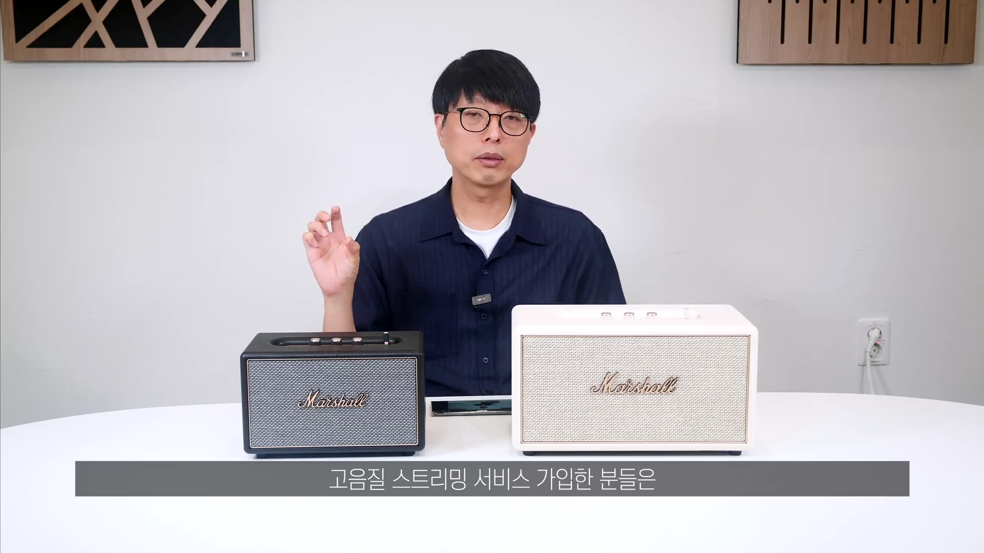 흰색 원형 테이블 위에 검은색과 크림색의 마샬 스피커 두 대가 놓여 있고, 뒤에서 안경을 쓴 남성이 손짓하며 설명하고 있는 스튜디오 인터뷰 영상 화면입니다.