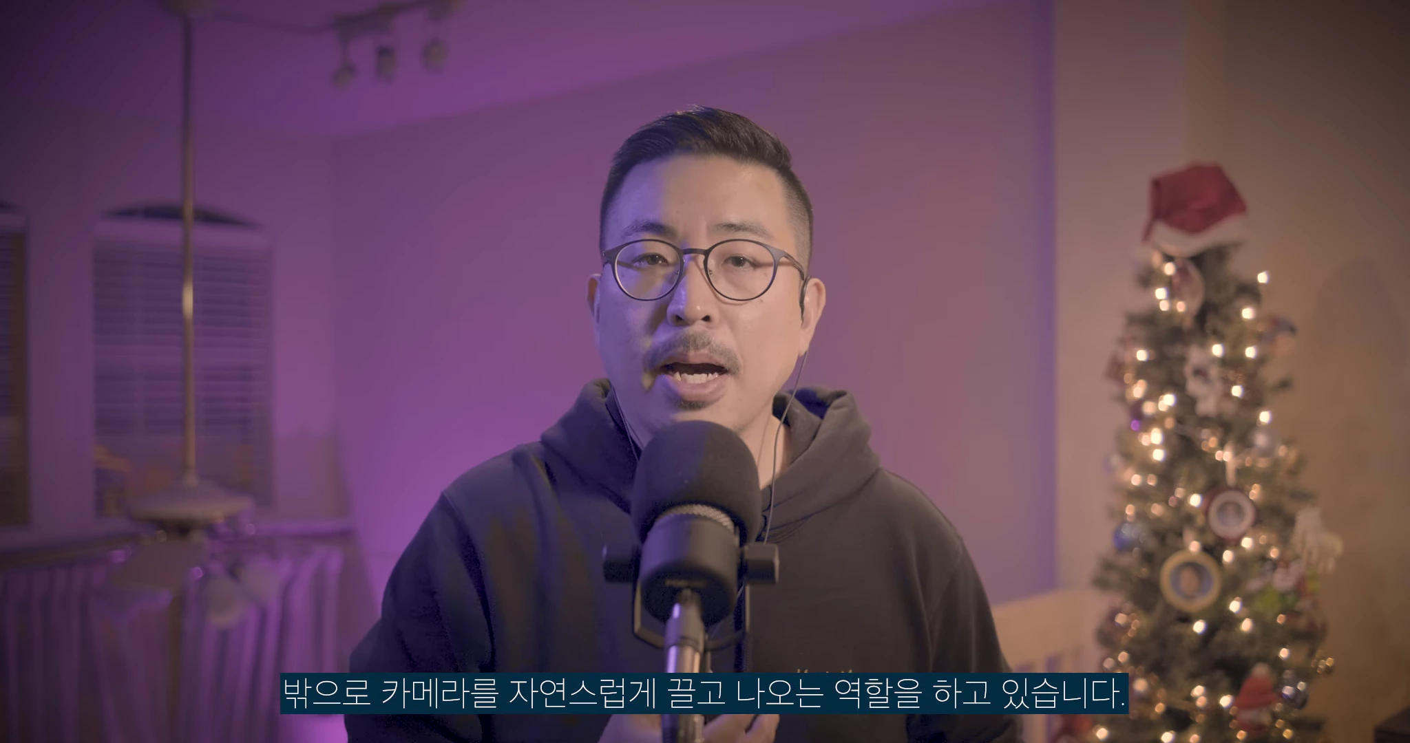 검은색 후드티를 입고 안경을 쓴 남성이 마이크 앞에서 이야기하고 있으며, 배경에는 불이 켜진 크리스마스트리가 보입니다.