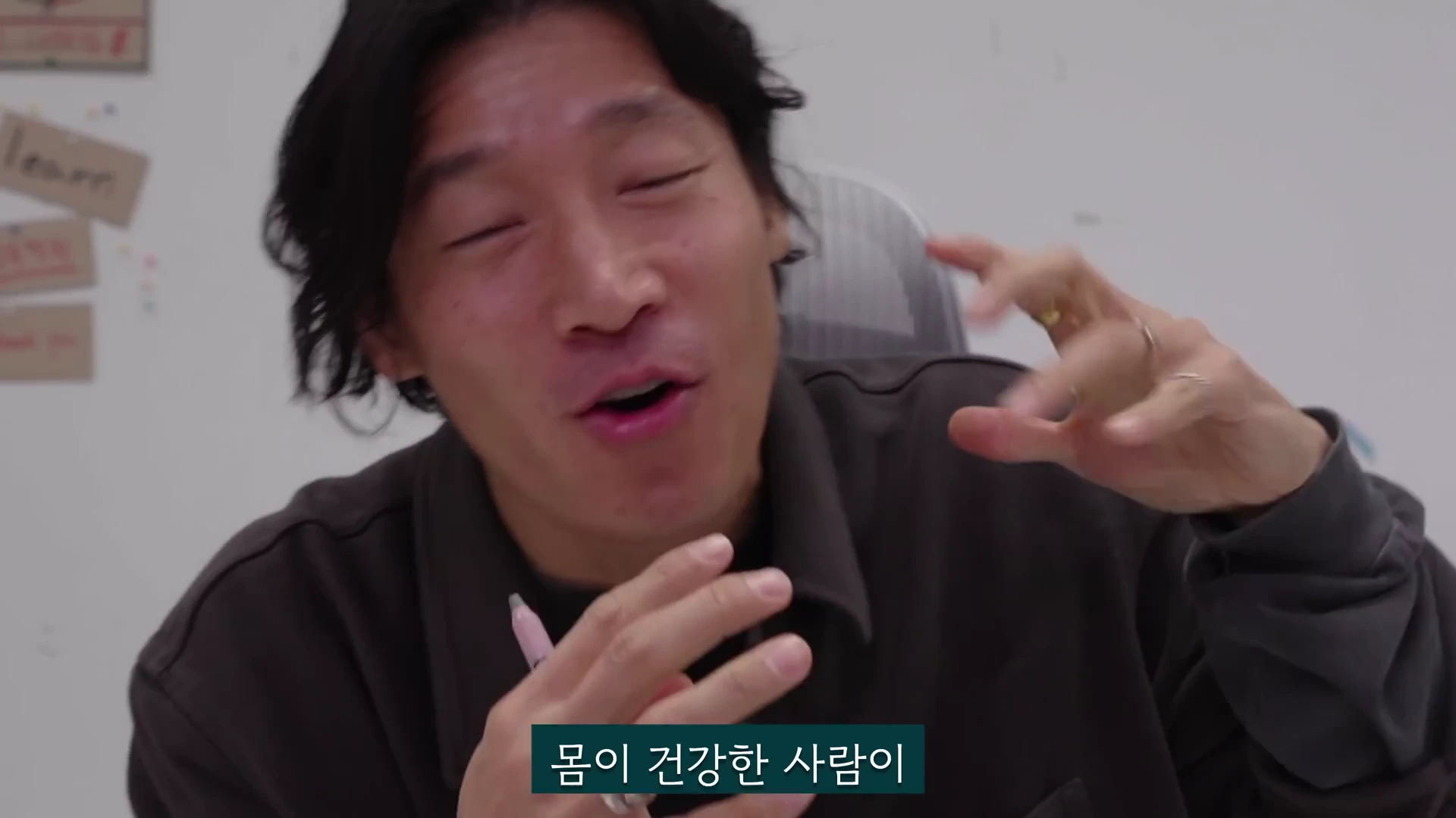 흰색 벽을 배경으로 어두운색 상의를 입은 남성이 손짓하며 이야기하고 있는 모습.