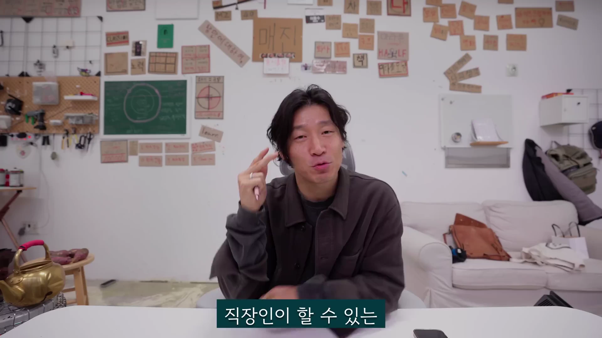 갈색 셔츠를 입은 남성이 방 안에서 책상에 앉아 카메라를 보며 손가락으로 제스처를 취하고 있다. 배경의 벽면에는 다양한 메모가 붙은 종이들이 여러 개 부착되어 있고, 왼편에는 초록색 칠판과 작업 도구들이 놓여 있다.