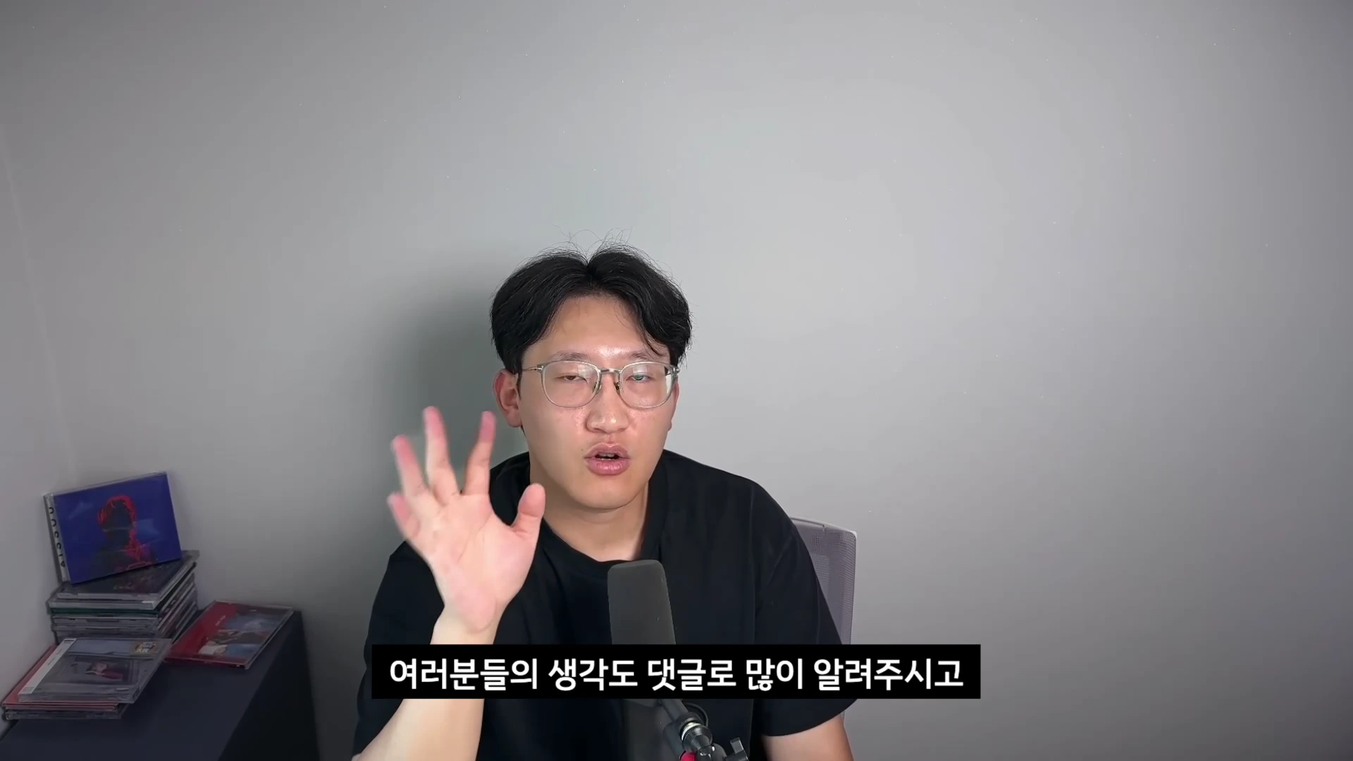 검은색 티셔츠를 입고 안경을 쓴 남성이 마이크 앞에 앉아 손을 흔들며 카메라를 향해 말하고 있는 모습