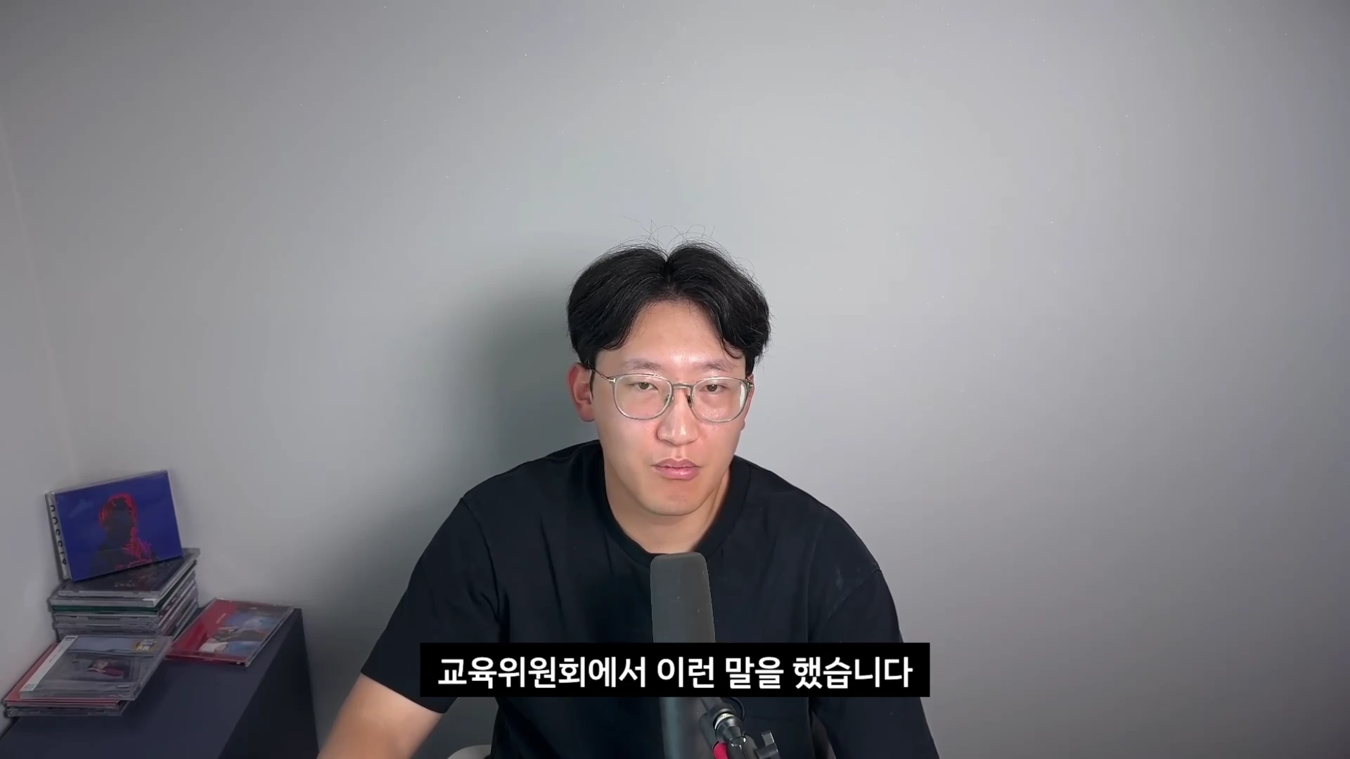검은색 티셔츠를 입은 안경 쓴 남성이 마이크 앞에서 이야기하고 있으며, 화면 하단에는 '교육위원회에서 이런 말을 했습니다'라는 한국어 자막이 표시되어 있습니다.