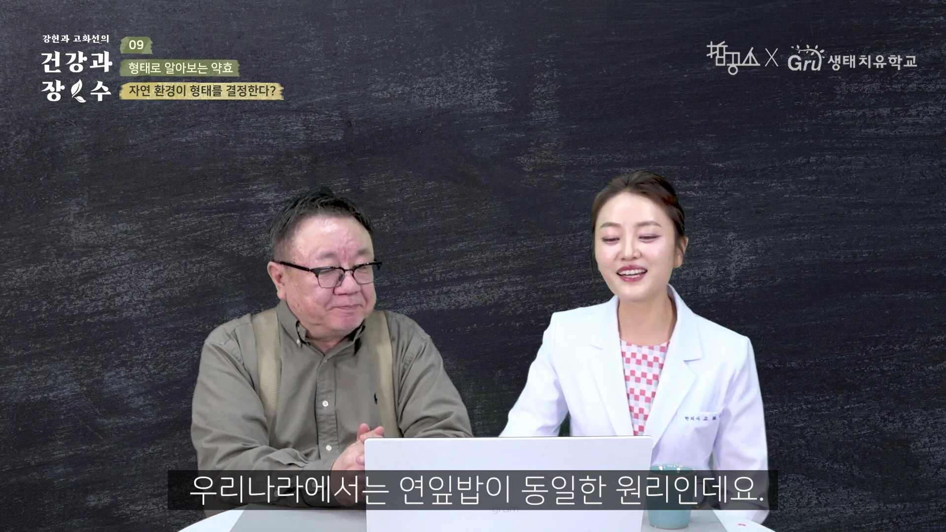 검은 배경의 스튜디오에서 중년 남성과 흰 의사 가운을 입은 여성이 테이블에 앉아 노트북 화면을 보며 대화를 나누고 있다.