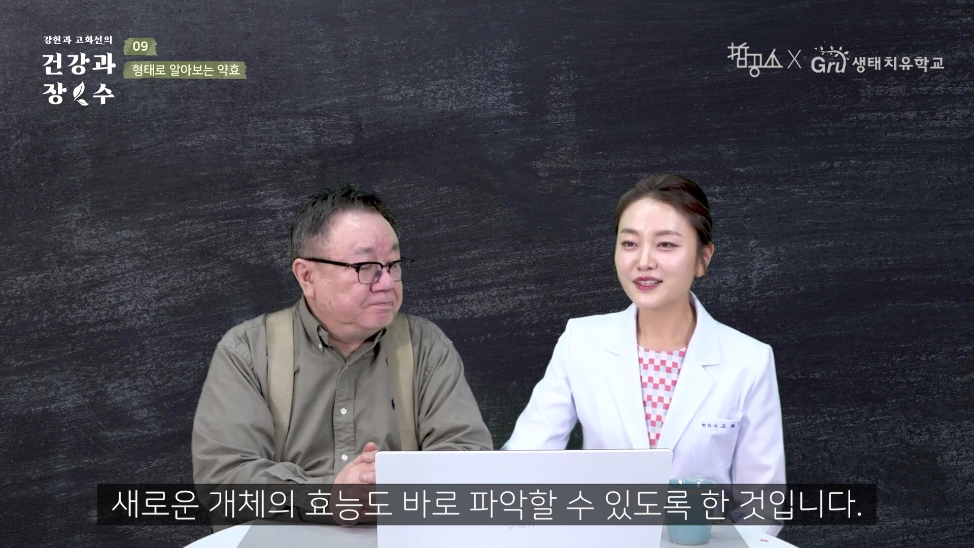 칠판 같은 배경 앞에서 안경을 쓴 중년 남성과 흰 가운을 입은 여성이 테이블에 앉아 대화하고 있는 모습이 담긴 영상 캡처 화면입니다.
