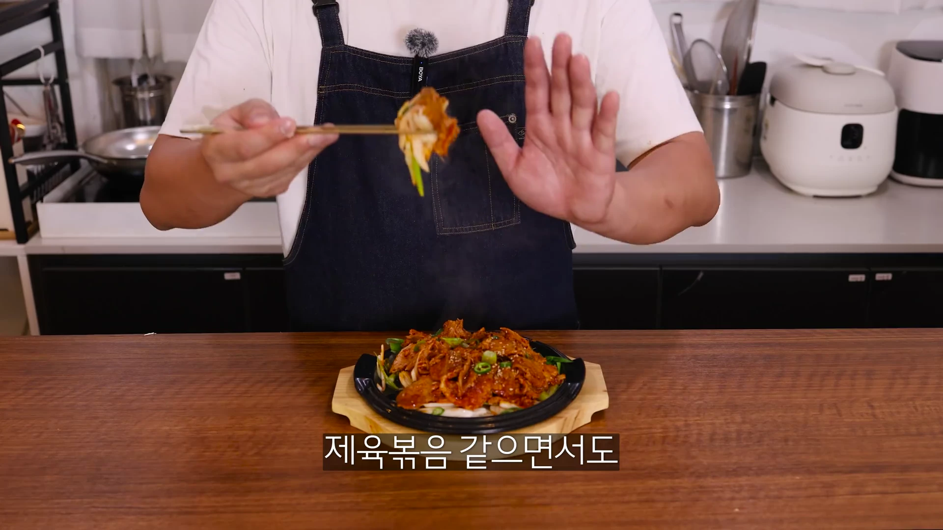 앞치마를 두른 사람이 젓가락으로 매콤한 양념에 볶은 고기를 들어 올리고 있는 모습이며, 앞쪽에는 철판에 담긴 요리가 놓여 있다.