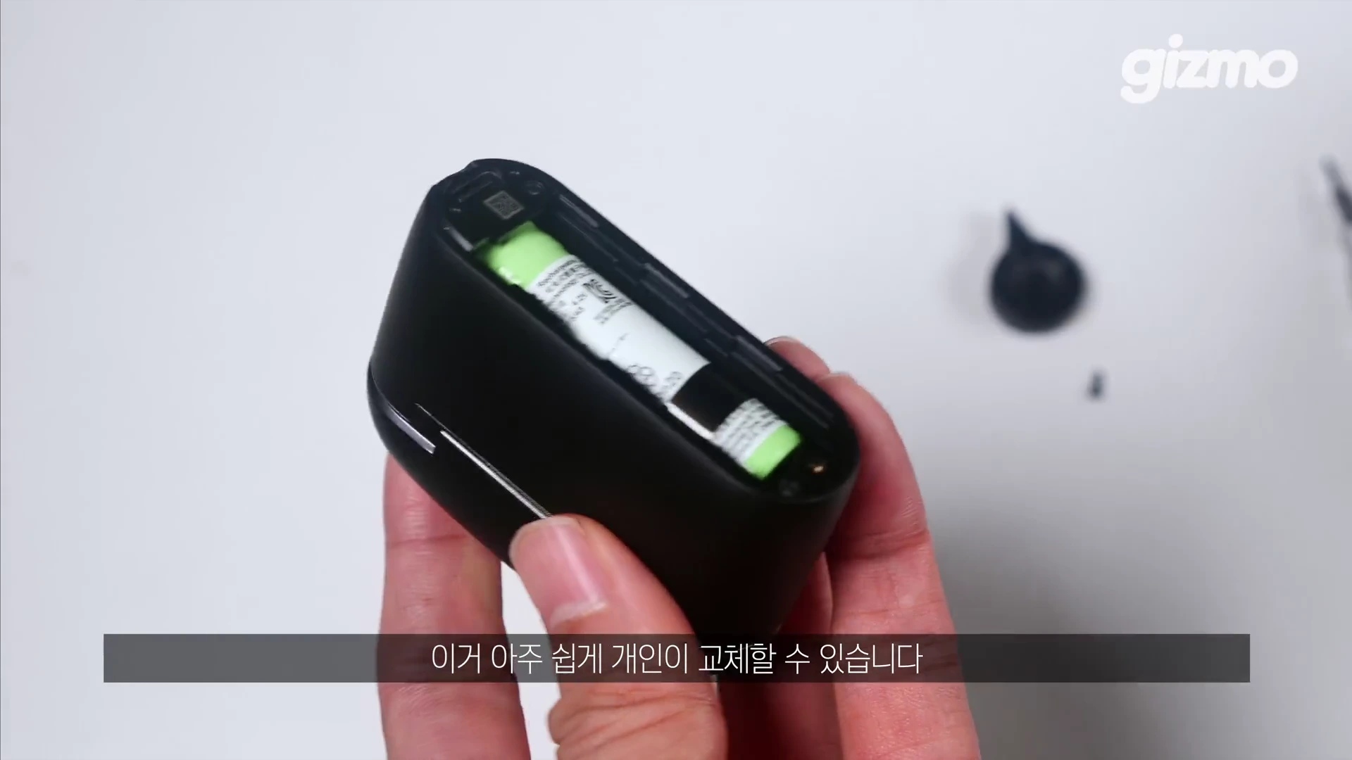 한 손으로 무선 이어폰 충전 케이스의 하단 덮개를 열어 내부의 원통형 배터리가 드러난 모습을 보여주는 클로즈업 영상 캡처 화면입니다.