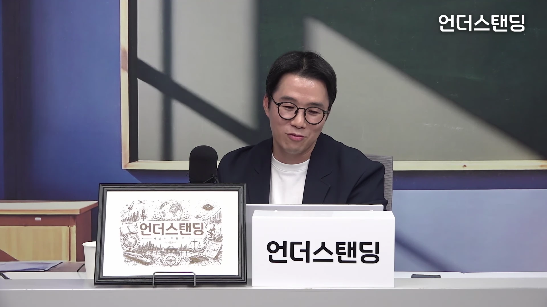 안경을 쓴 남성이 스튜디오 테이블에 앉아 카메라를 보며 이야기하고 있다.