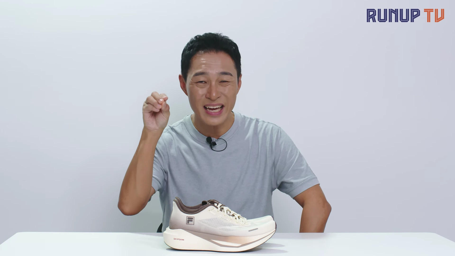 흰 배경의 스튜디오에서 티셔츠를 입은 남성이 테이블 위에 놓인 베이지색 러닝화를 가리키며 손짓하고 있다.