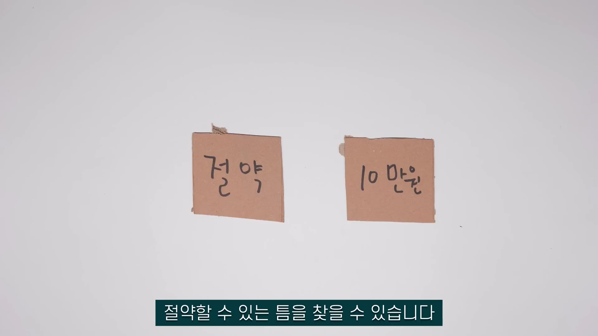 흰색 배경 위에 '절약'과 '10만 원'이라고 적힌 두 개의 작은 정사각형 갈색 종이 조각이 나란히 놓여 있고, 하단에는 검은색 배경에 흰 글씨로 '절약할 수 있는 틈을 찾을 수 있습니다'라는 자막이 표시되어 있다.
