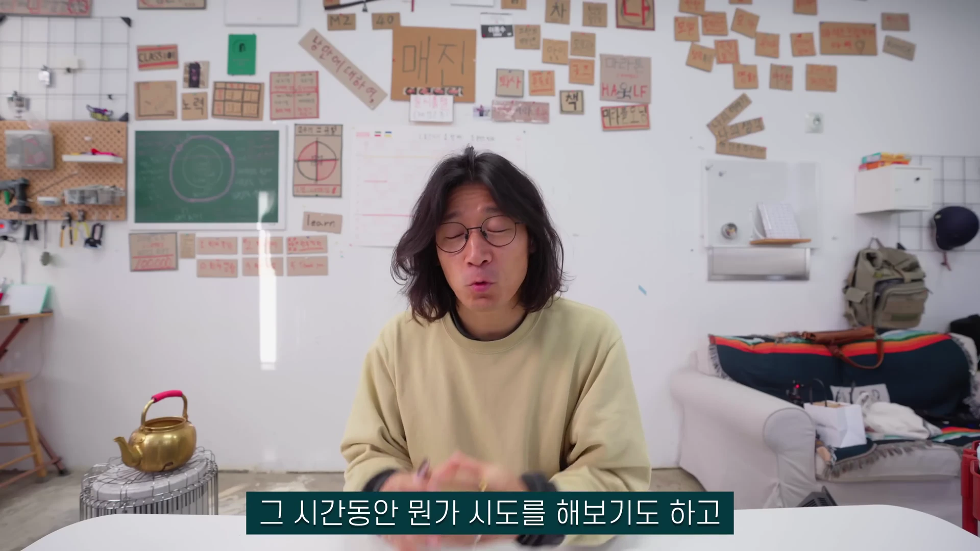 안경을 쓴 긴 머리의 남성이 밝은 실내 공간에서 이야기를 나누고 있으며, 배경 벽면에는 다양한 메모지가 붙어 있다.