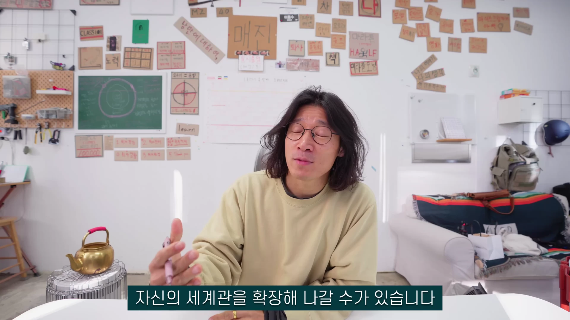 긴 머리에 안경을 쓴 남성이 베이지색 상의를 입고 방 안에서 손동작을 하며 이야기를 나누고 있다. 배경 벽면에는 다양한 메모와 글귀가 적힌 종이들이 붙어 있다.