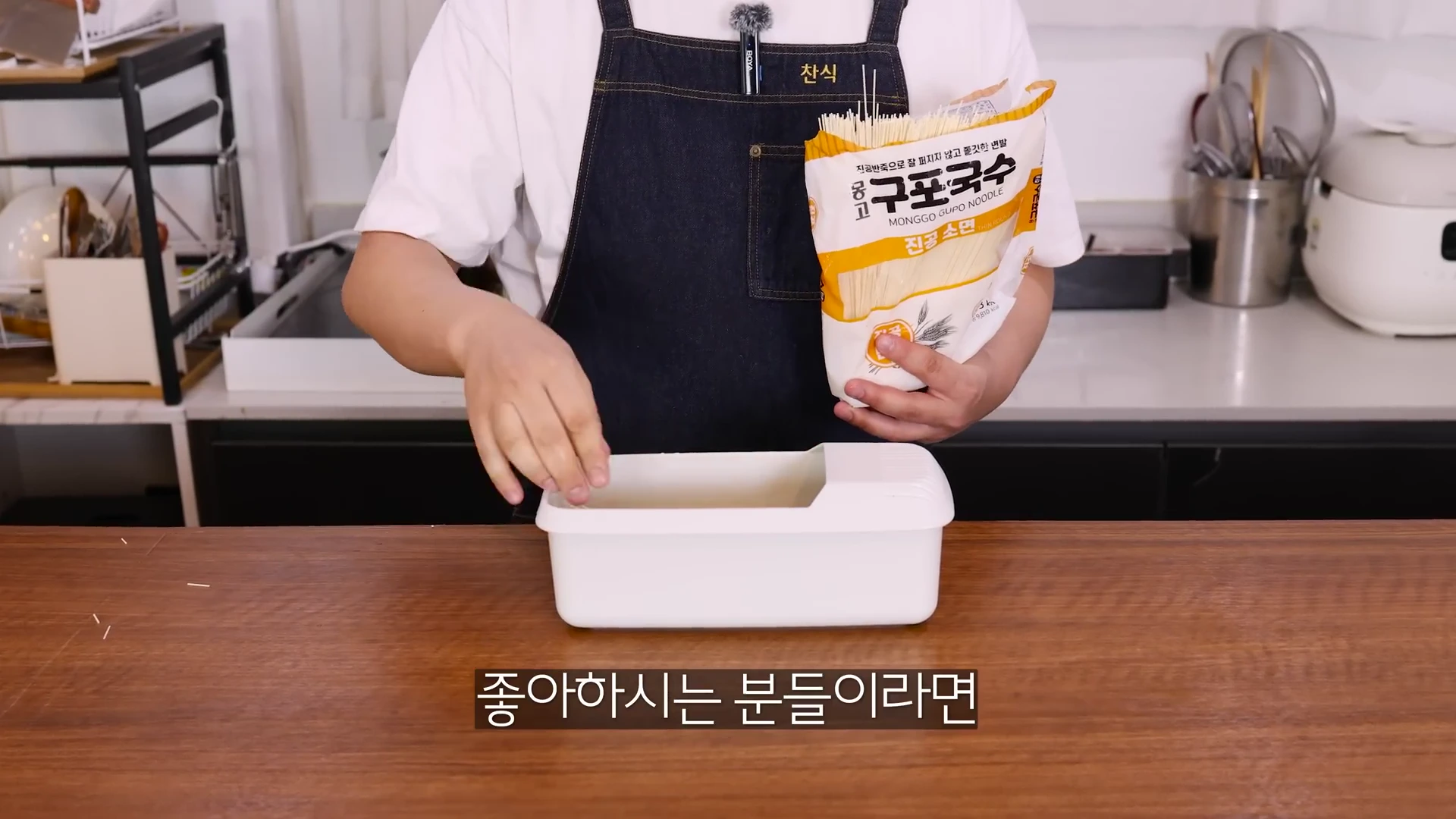 앞치마를 두른 사람이 손에 국수 봉지를 들고 흰색 직사각형 모양의 파스타 쿠커 안에 면을 넣고 있는 모습입니다.