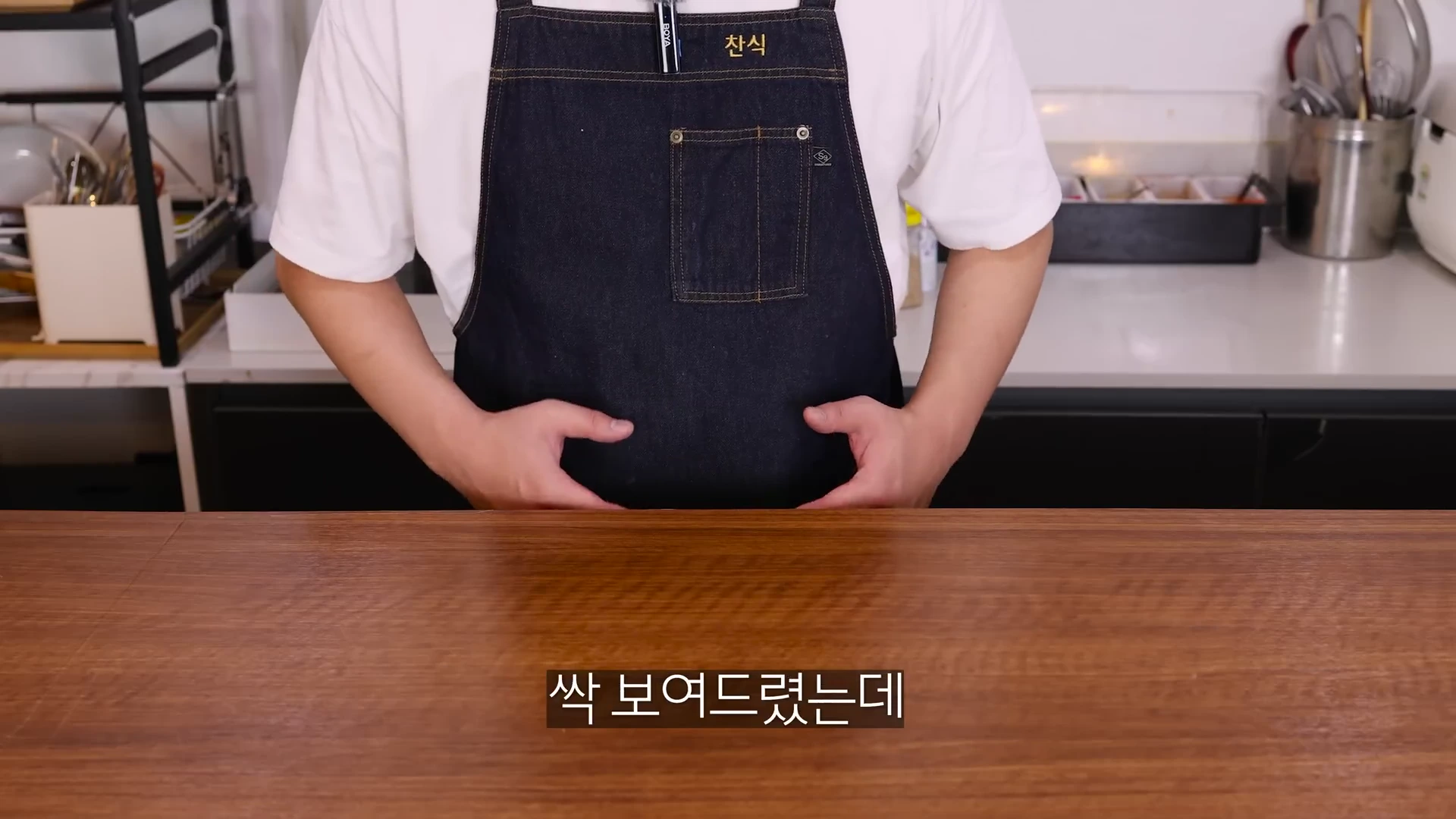 앞치마를 입은 한 남성이 나무 식탁 뒤에 서 있는 모습