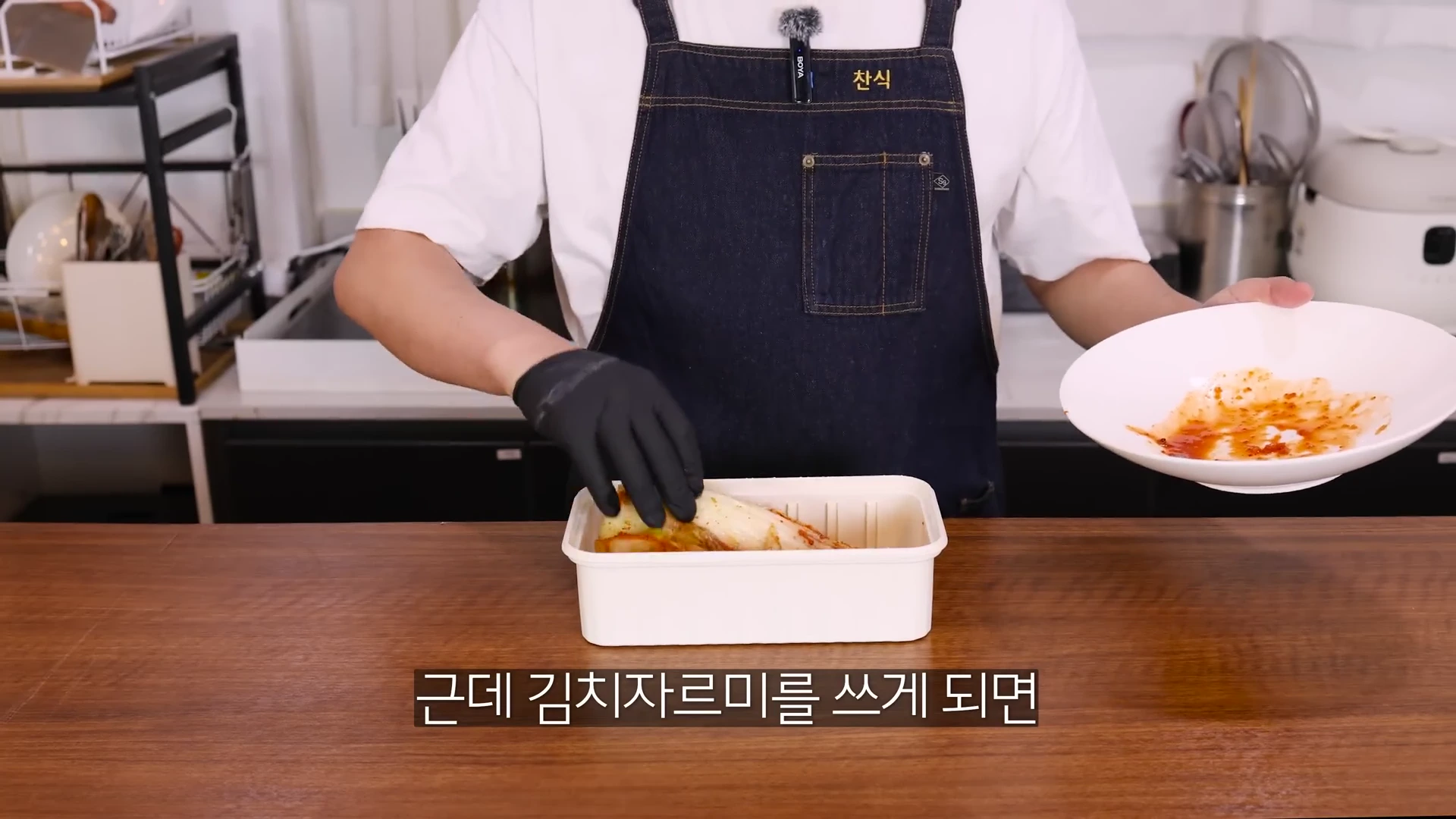 흰색 티셔츠에 앞치마를 두른 사람이 검은색 장갑을 끼고 흰색 김치통을 다루고 있으며, 자막으로 '근데 김치자르미를 쓰게 되면'이라고 적혀 있는 영상 프레임입니다.