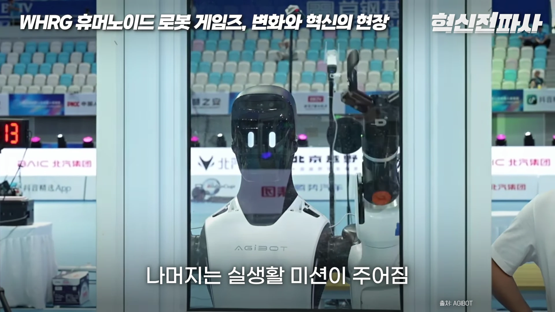 베이징의 경기장에서 흰색과 검은색으로 이루어진 휴머노이드 로봇이 한쪽 팔을 들어 올린 채 서 있고, 화면 하단에는 '나머지는 실생활 미션이 주어짐'이라는 자막이 적혀 있다.