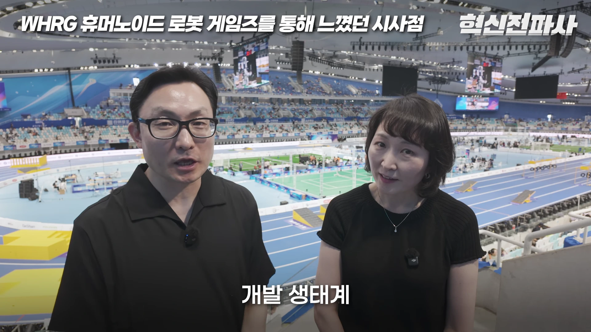 실내 스포츠 경기장 관람석에서 두 명의 남녀가 카메라를 정면으로 바라보며 대화하고 있고, 배경으로는 넓은 경기장 트랙과 로봇 관련 경기가 진행 중인 모습이 흐릿하게 보입니다.