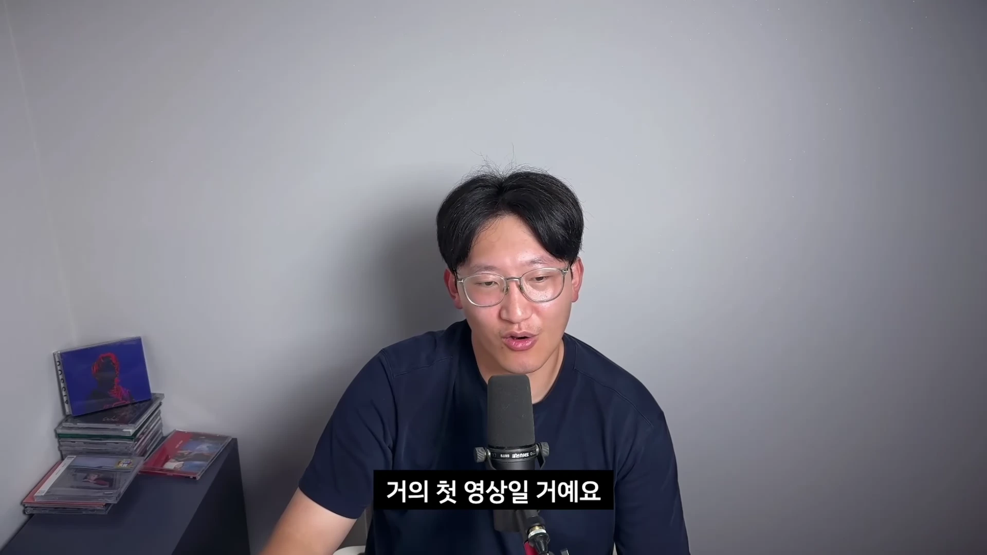 안경을 쓴 한 남성이 회색 배경 앞에서 마이크를 앞에 두고 이야기를 하고 있으며 왼쪽에는 앨범들이 놓여 있다.