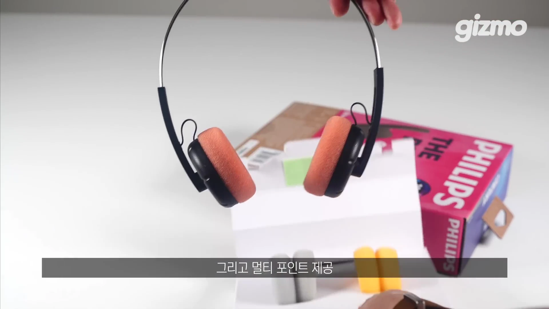 주황색 이어 패드가 달린 검은색 레트로 스타일 헤드폰을 손으로 들어 올리고 있으며, 배경에는 제품 포장 상자와 교체용 이어 패드가 놓여 있습니다. 화면 하단에는 멀티 포인트 기능을 설명하는 한글 자막이 떠 있습니다.