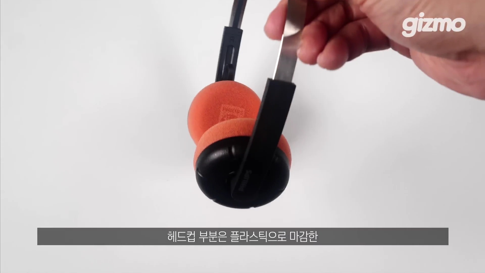 손으로 들고 있는 온이어 방식의 검은색 무선 헤드폰으로, 오렌지색 스펀지 이어패드와 스테인리스 스틸 헤드밴드 구조가 강조되어 보임