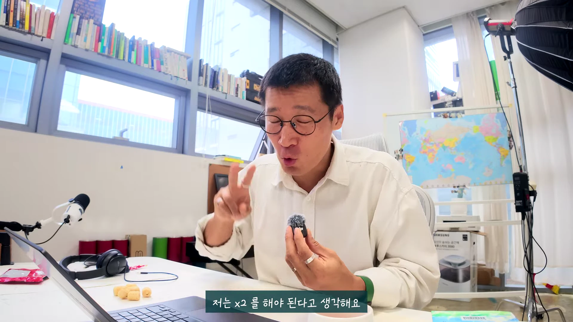 안경을 쓴 남성이 밝은 사무실 책상에 앉아 작은 무선 마이크를 손에 쥐고 손가락으로 숫자 2를 표시하며 이야기하고 있다.