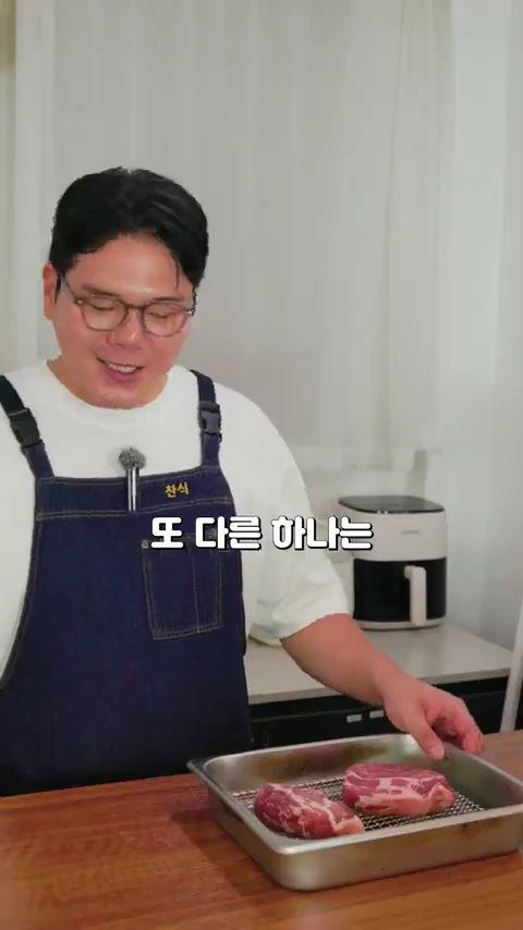 안경을 쓴 남성이 앞치마를 입고 테이블 위에 놓인 두 덩이의 돼지고기를 가리키고 있는 모습
