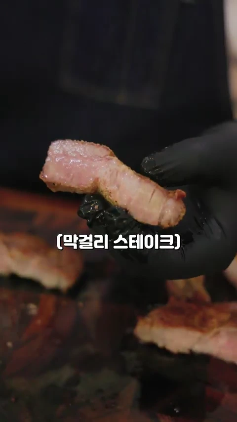 검은색 위생 장갑을 낀 손이 잘 구워진 돼지고기 한 점을 들어 보이고 있으며, 화면 하단에는 막걸리 스테이크라는 텍스트가 표시되어 있습니다.