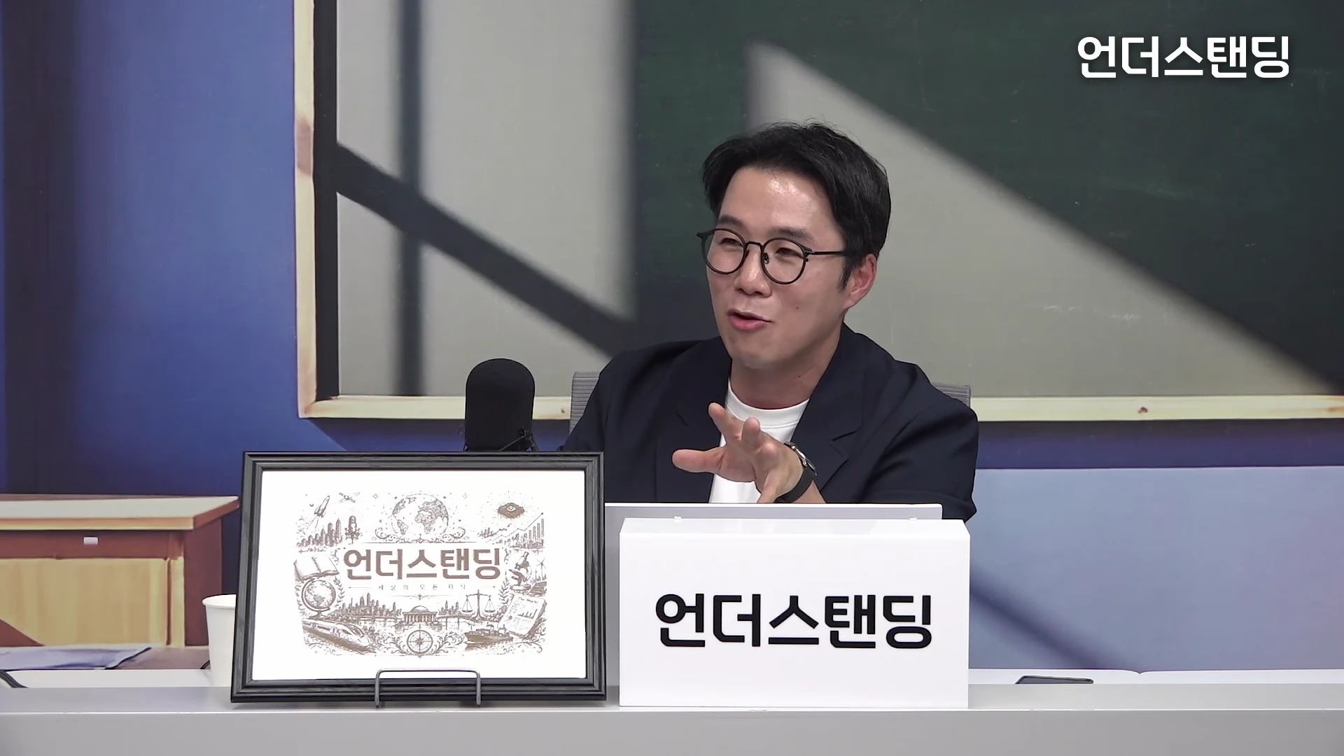 안경을 쓴 남성이 스튜디오 책상에 앉아 손짓하며 대화하는 모습. 앞에는 로고가 그려진 액자와 흰색 명패가 놓여 있다.