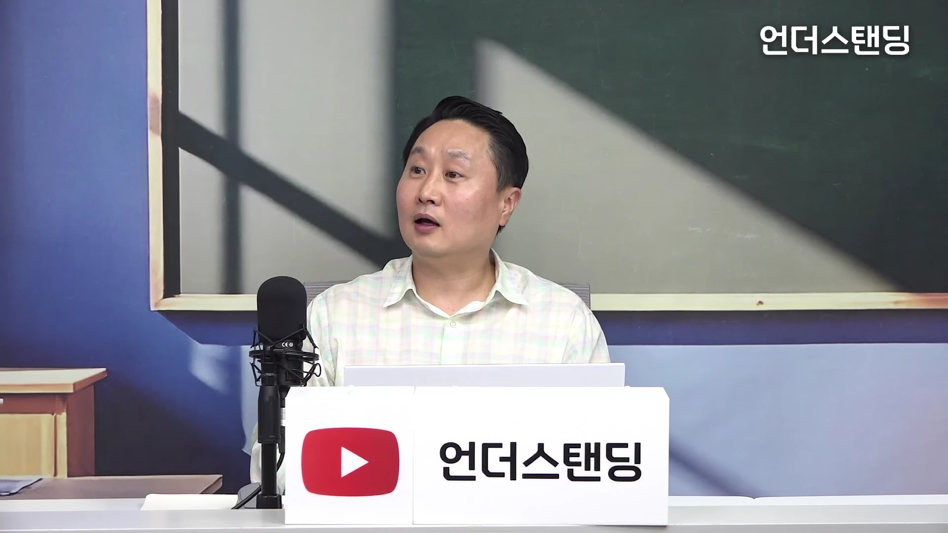 한 남성이 스튜디오 책상 앞에 앉아 앞에 있는 마이크를 보고 이야기하고 있으며, 앞쪽에는 로고가 새겨진 흰색 표지판이 놓여 있습니다.