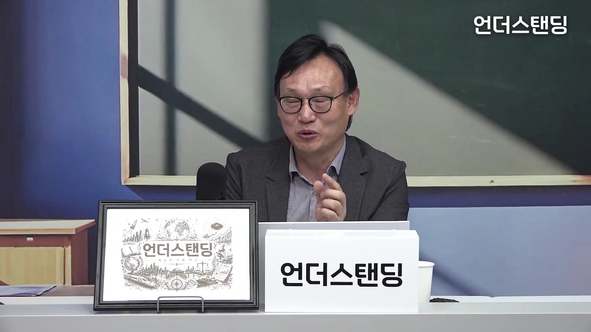 안경을 쓴 중년 남성이 책상 앞에 앉아 이야기하는 모습으로, 앞에는 로고가 그려진 액자와 하얀색 안내판이 놓여 있다.