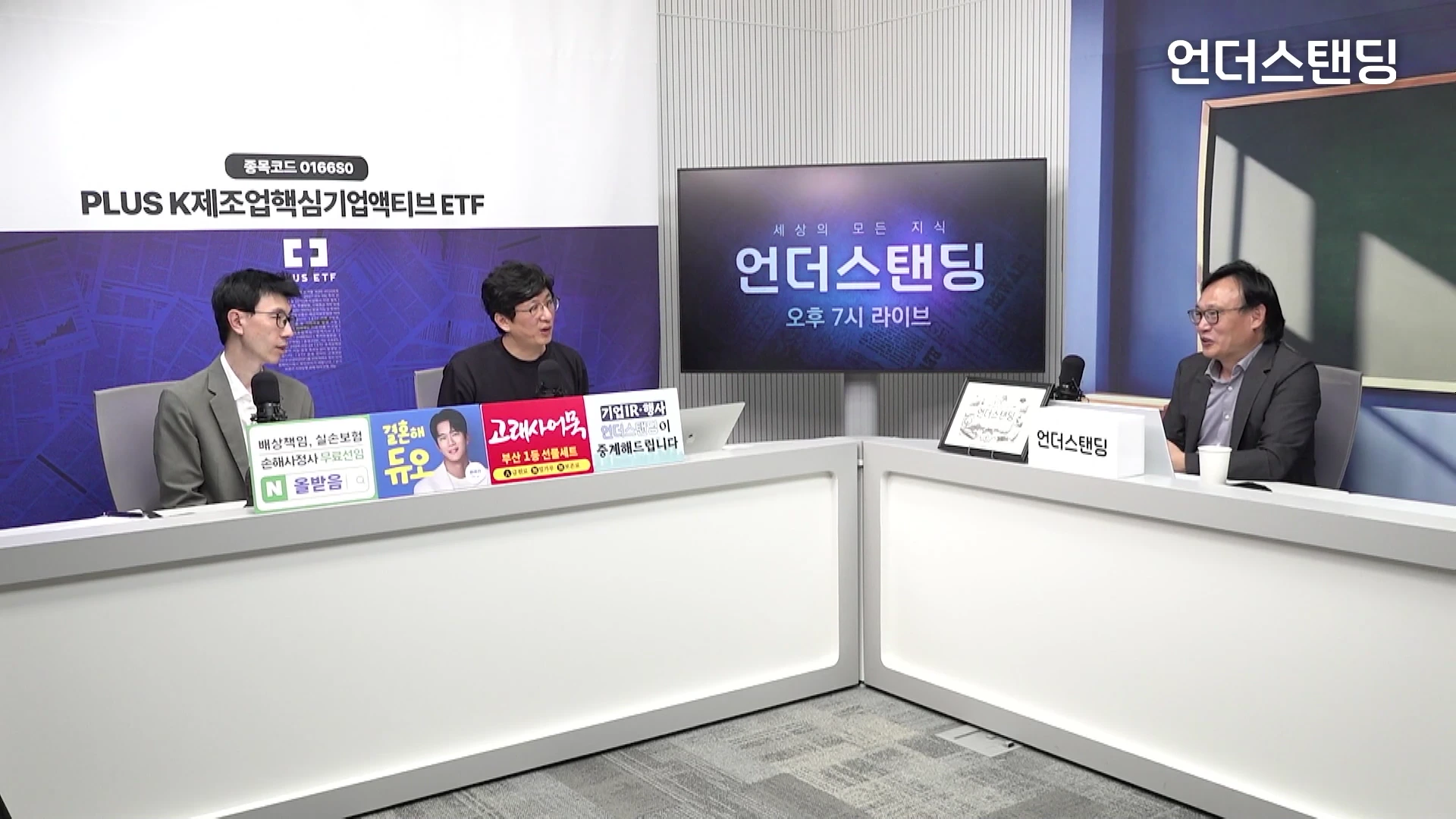 세 명의 남성이 스튜디오 책상에 앉아 대담을 나누고 있는 모습으로, 배경에는 로고가 표시된 대형 화면과 광고판들이 배치되어 있다.
