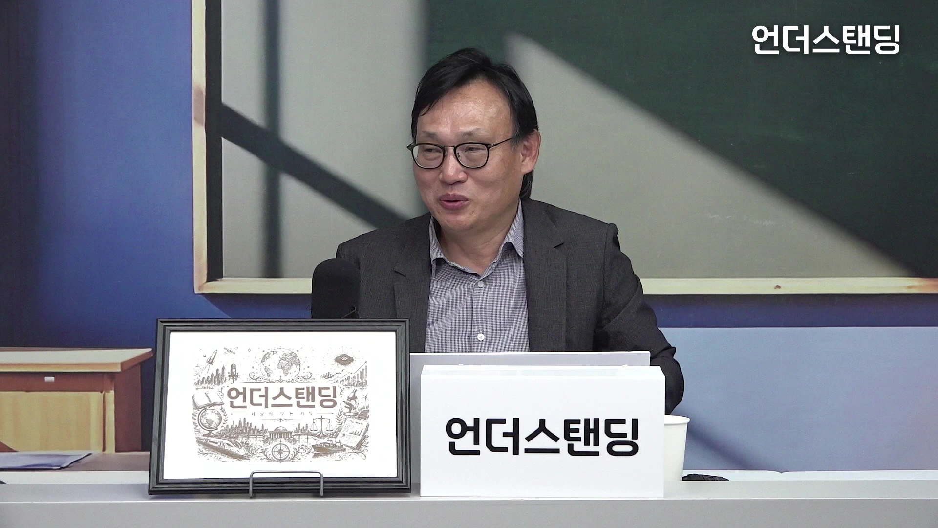 안경을 쓴 중년 남성이 스튜디오 데스크에 앉아 인터뷰를 진행하고 있으며, 앞에는 로고가 그려진 액자와 명패가 놓여 있습니다.