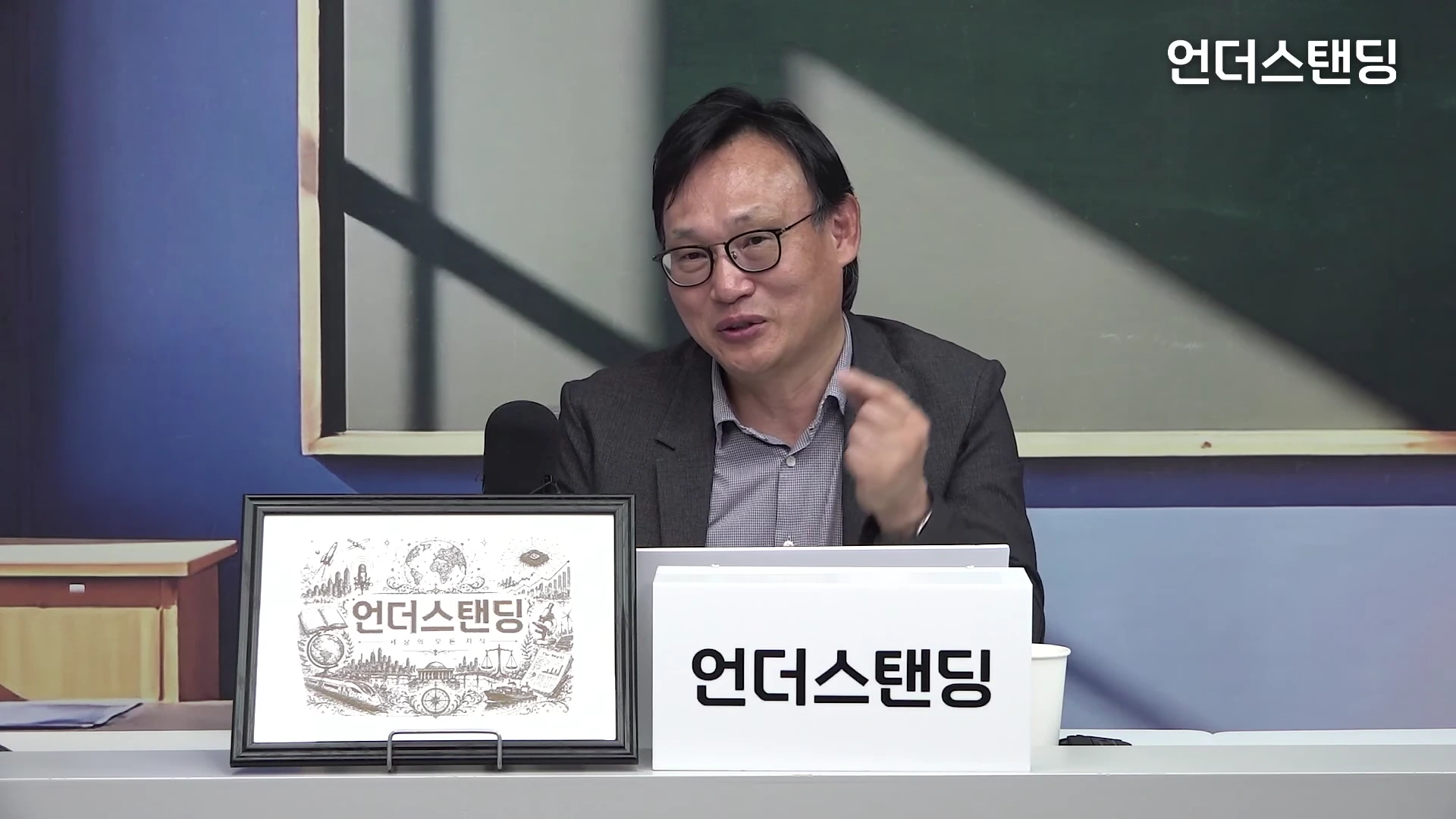 안경을 쓴 중년 남성이 스튜디오 테이블 앞에 앉아 손가락을 들어 올리며 설명하고 있는 모습으로, 앞에는 로고가 그려진 액자와 명패가 놓여 있습니다.