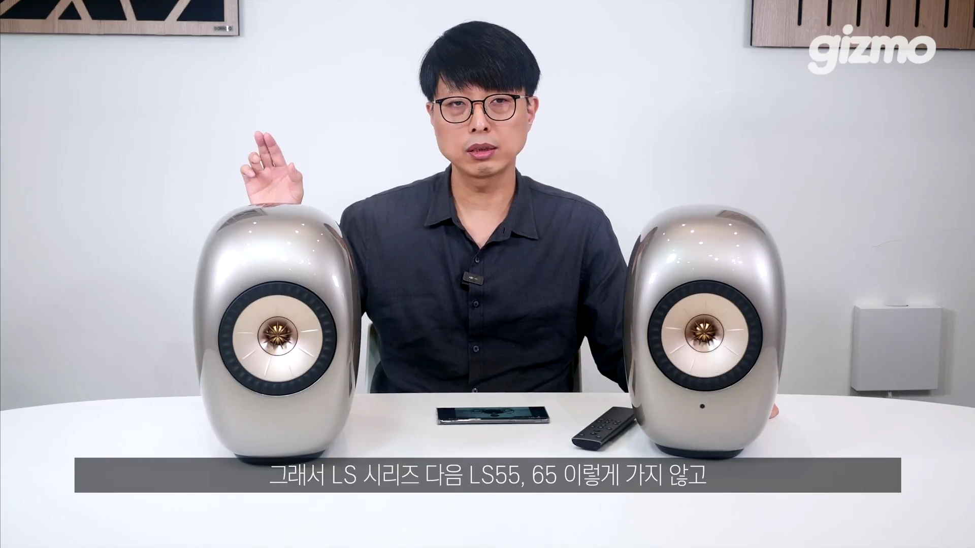 한 남성이 양쪽에 은색 스피커를 두고 책상 앞에 앉아 설명하고 있는 모습