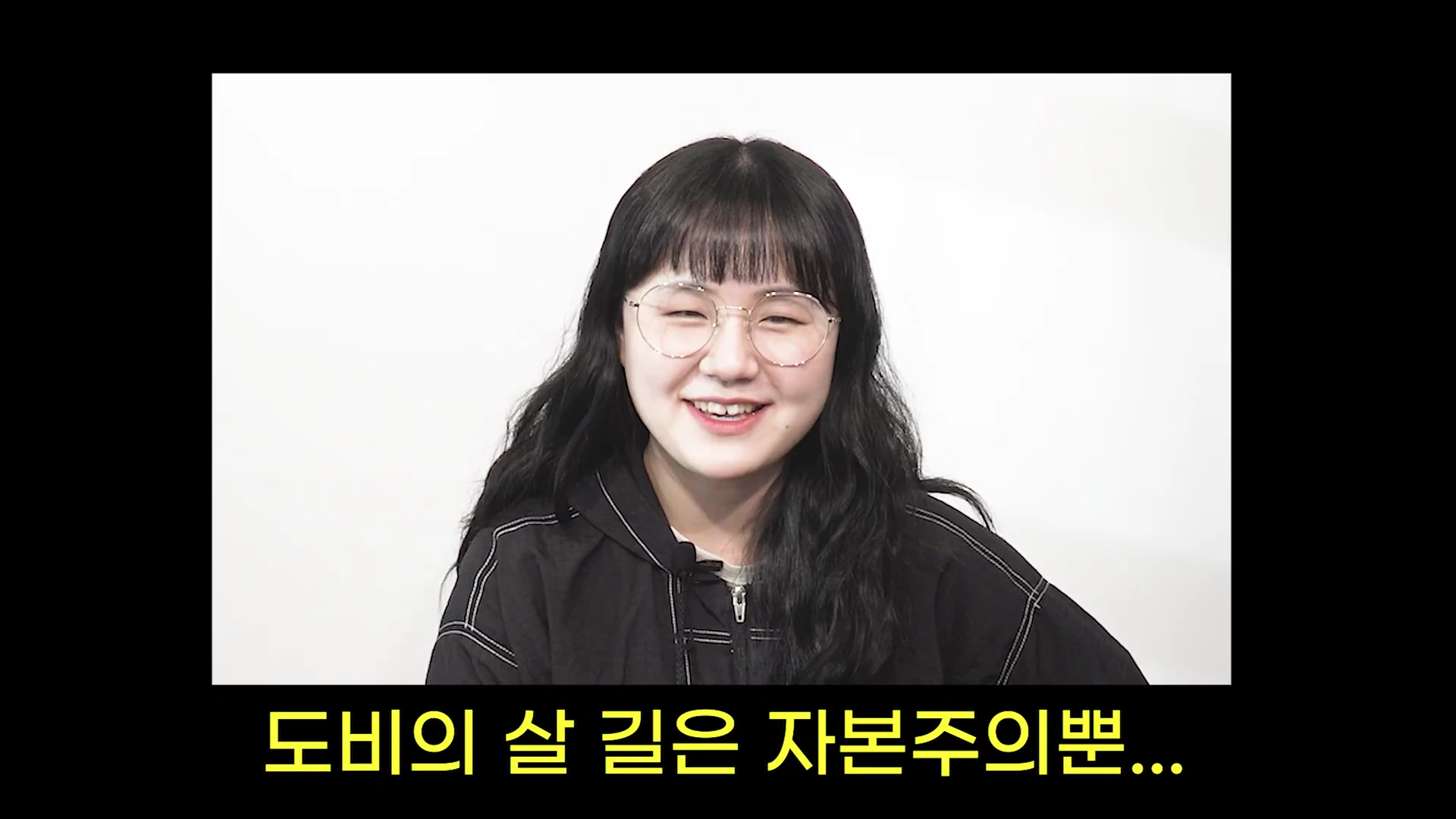 흰색 배경 앞에 안경을 쓴 여성이 밝게 웃고 있으며, 화면 하단에는 노란색 자막으로 '도비의 살 길은 자본주의뿐...'이라는 문구가 적혀 있습니다.
