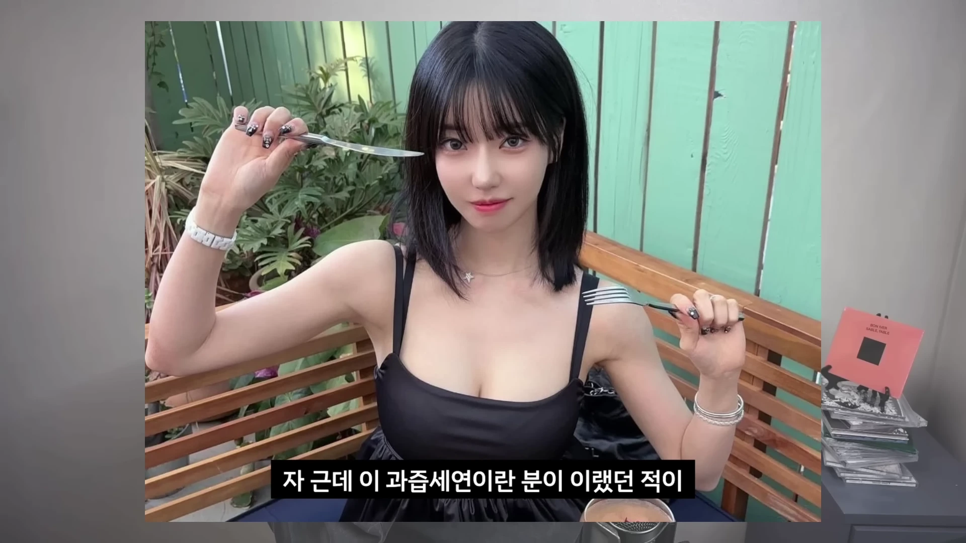 긴 머리의 젊은 여성이 야외 테이블에 앉아 나이프와 포크를 들고 카메라를 응시하는 모습이 담긴 유튜브 영상 캡처 화면